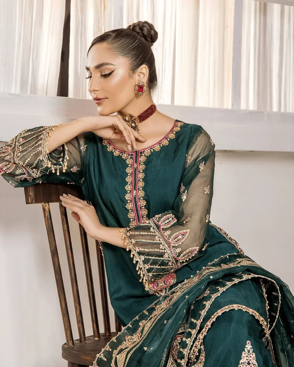 Woman in a teal embroidered traditional outfit sitting on a wooden chair.